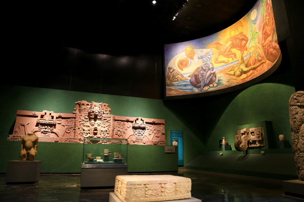 Ausstellungsraum im Anthropologischen Museum von Mexiko-Stadt, der verschiedene Kunstwerke zeigt, darunter Skulpturen, Tischobjekte, eine Texttafel, eine Wandmalerei und Deckenbeleuchtung.