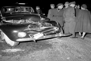 Ein Schwarz-Weiß-Bild eines demolierten Autos am Straßenrand mit eingedellter Motorhaube und zersplitterten Fenstern, umgeben von einer Gruppe besorgter Menschen.