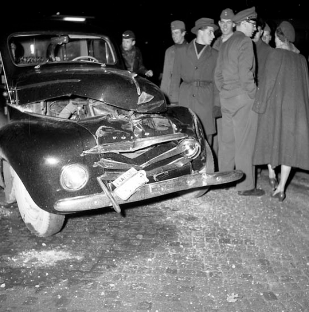 Ein Schwarz-Weiß-Bild eines demolierten Autos am Straßenrand mit eingedellter Motorhaube und zersplitterten Fenstern, umgeben von einer Gruppe besorgter Menschen.