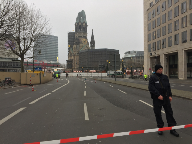 Ein Polizist in schwarzer Uniform und Mütze steht in einer Berliner Straße umgeben von Absperrbaken, Fahrrädern, Laternen, Bäumen und Gebäuden, mit einem Kirchturm und einem klaren blauen Himmel im Hintergrund.