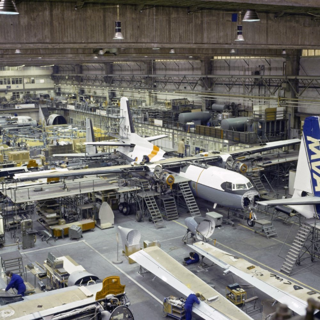 Ein großer Hangar voller Flugzeuge auf der Montage, Arbeiter, Leitern und diverse Gegenstände auf dem Boden, mit Lampen an der Decke und einer Wand im Hintergrund.