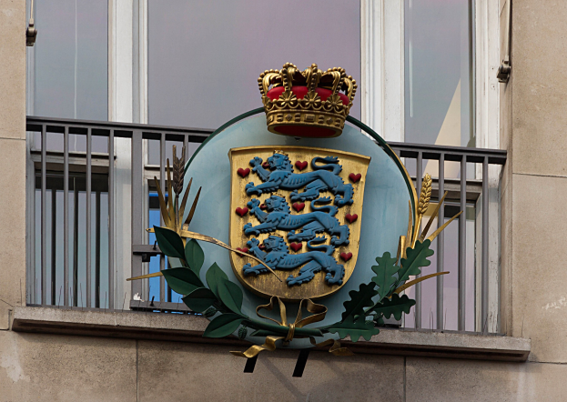 Das Wappen der britischen Königsfamilie, das auf der Außenseite eines Gebäudes dargestellt ist, begleitet von einem Kronen- und Blätterlogo, mit Fenstern und Metallgittern im Hintergrund.