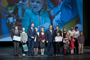 Eine Gruppe von Menschen auf einer Bühne mit Taschen und Urkunden, im Hintergrund ein Bildschirm mit einem Kind und anderen, was auf ein Charity-Event hinweist.