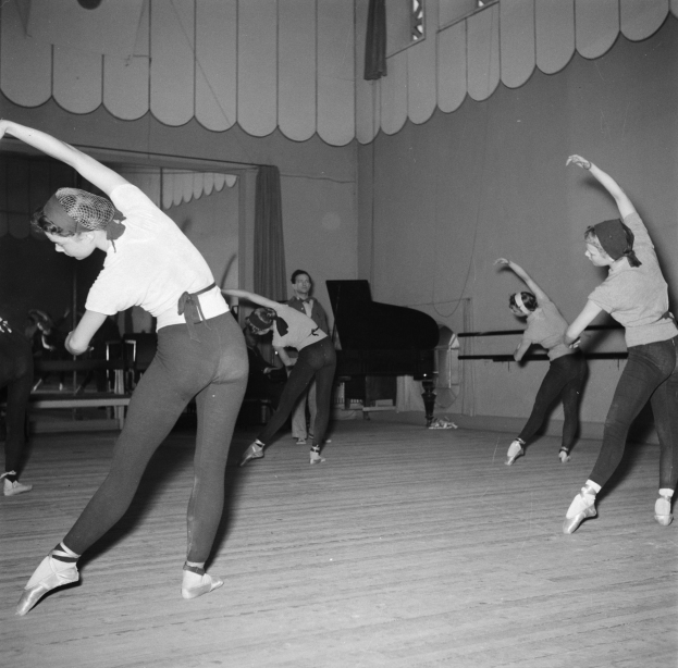 Eine Gruppe junger Frauen tanzt anmutig in einem schwarzen und weißen Studio, mit einem Piano im Hintergrund und einer Wand dahinter.