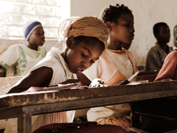 Eine Gruppe von Kindern sitzt an Tischen in einem Klassenzimmer, mit Büchern und Taschen auf den Bänken vor ihnen und einer Wand mit einem Fenster im Hintergrund.