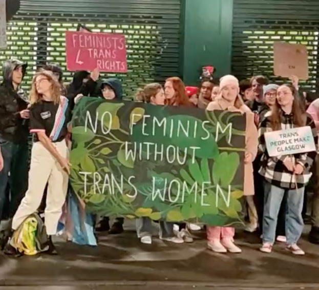 Eine Gruppe von Menschen mit einem Transparent, auf dem "Kein Feminismus ohne Transfrauen" steht, und verschiedenen Schildern, die in einer öffentlichen Fläche stehen.