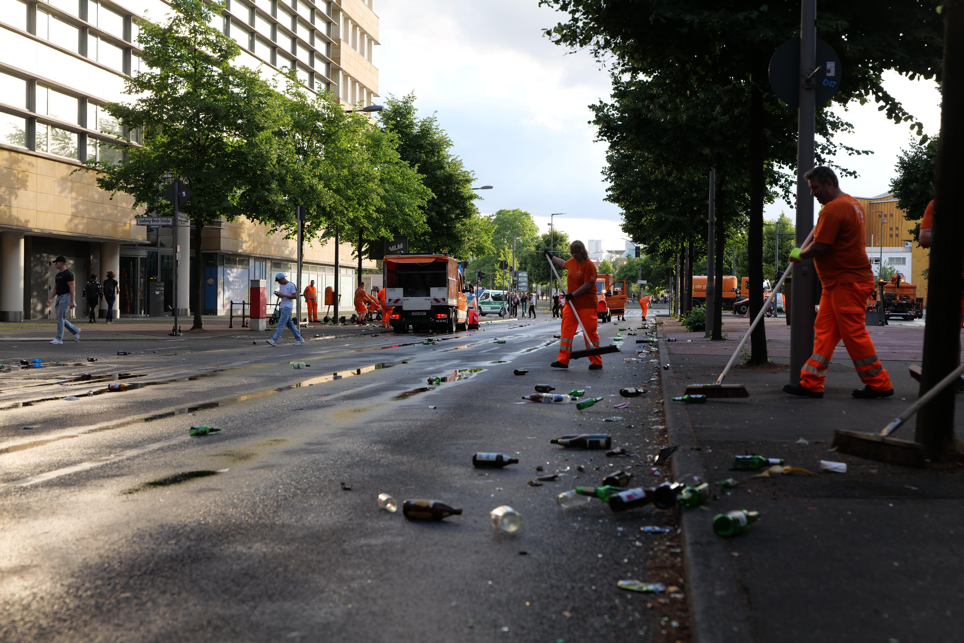 Eine Gruppe von Menschen in orangen Uniformen sammelt Müll von einer Straße, die mit Flaschen und Schutt übersät ist, mit Bäumen, Polen und Fahrzeugen im Hintergrund unter einem bewölkten Himmel.