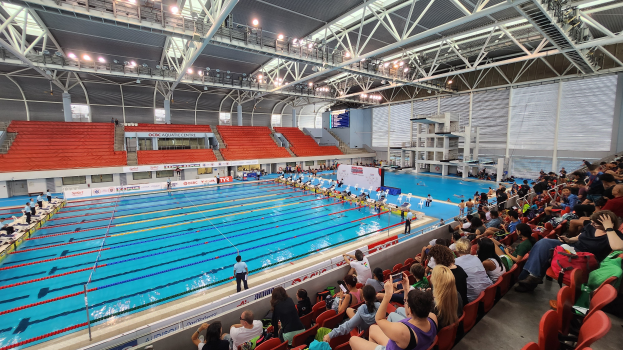 Ein großes Hallenbad mit vielen Menschen auf Stühlen drumherum und einigen im Wasser Stehenden sowie Schildern im Hintergrund und einer Decke mit Lampen und Eisenstangen.