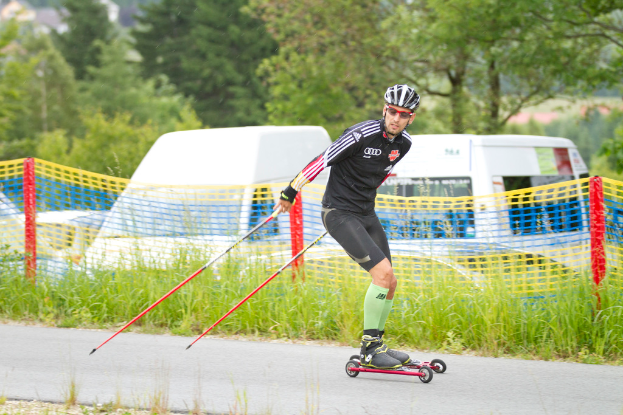 Eine Person fährt Skateboard auf einer Straße mit Gras, Netzen, Fahrzeugen und Bäumen im Hintergrund.