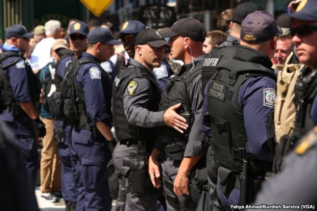Gruppe von Polizisten in Uniform auf einer Straße stehend, einige halten Waffen, mit Gebäuden und Beschriftungen im Hintergrund.