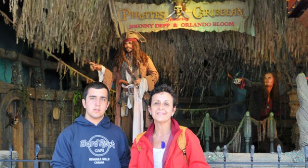Ein Mann und eine Frau stehen vor einem Piratenschiff mit einem Geländer am Boden, während Statuen in Kostümen, eine Fahne mit Text und Lichter im Hintergrund zu sehen sind.