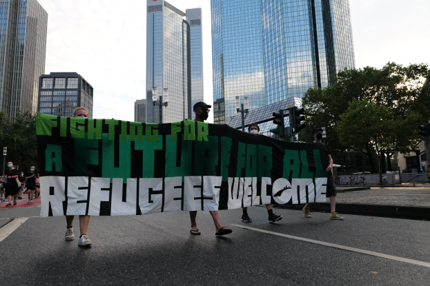 Eine Gruppe von Menschen marschiert auf einer von Bäumen gesäumten Straße, die eine Fahne schwenkt, auf der steht "Kämpfen für eine Zukunft für alle Flüchtlinge Willkommen", mit Gebäuden und Verkehrszeichen im Hintergrund unter einem bewölkten Himmel.