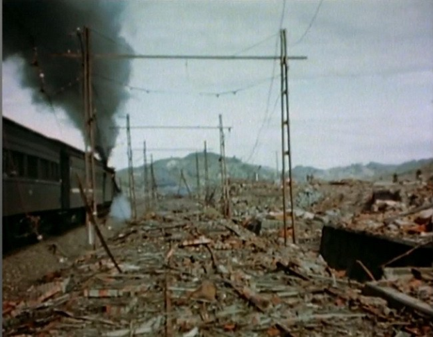 Ein Zug fährt durch ein beschädigtes Gebiet mit aufsteigendem Rauch, umgeben von Strommästen und -drähten, mit Höhen und einem klaren Himmel im Hintergrund und verstreutem Schutt auf dem Boden.