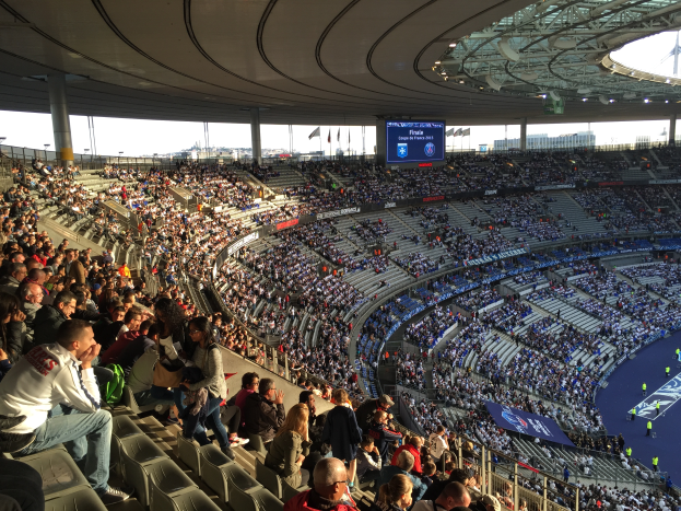Eine große Menschenmenge sitzt in einem Stadion und schaut ein Fußballspiel. Rechts im Bild befindet sich eine Bühne mit einigen Personen darauf. Im Hintergrund sind Fahnen, Stangen und ein Bildschirm zu sehen. Der Himmel ist oben im Bild zu erkennen, und das Stadion scheint die Allianz Arena in München, Deutschland, zu sein.