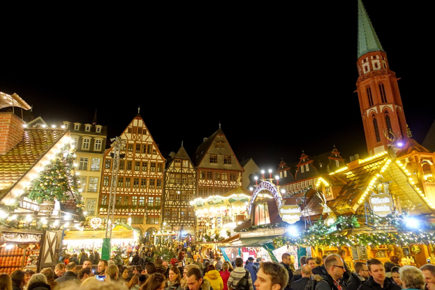 Ein geschäftiger Weihnachtsmarkt in Nürnberg, Deutschland mit Menschen um geschmückte Stände, festliche Beleuchtung, Gebäude mit Fenstern, ein Uhrturm und einen dunklen Himmel.