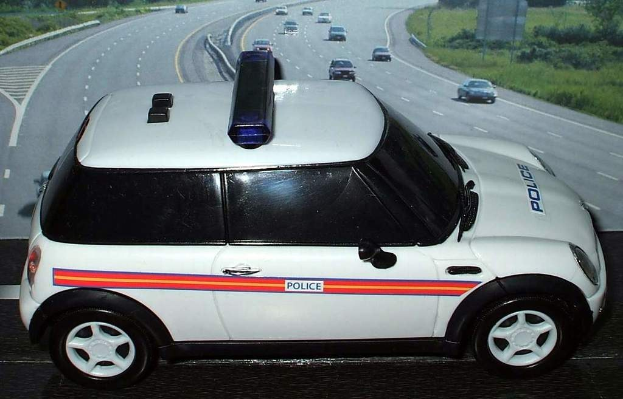Weißer Polizeiwagen vor bewegendem Verkehr auf einer Straße gesäumt von Gras und Bäumen außerhalb der Stadt.