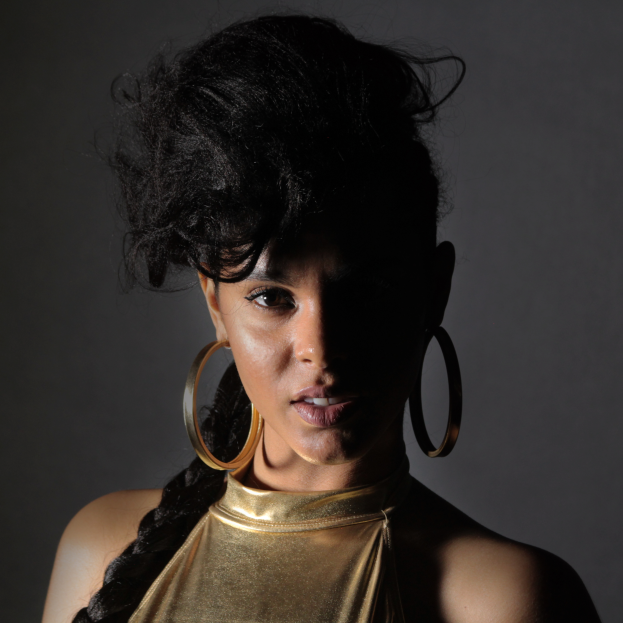 Eine Frau in einem goldenen Kleid und großen Creolen steht selbstbewusst vor einer Wand, ihre glatte Bob-Frisur ist ordentlich gestylt, sie trägt dezenten natürlichen Make-up mit pinkem Rouge und glänzenden Lippen und schaut direkt in die Kamera mit einem entschlossenen Gesichtsausdruck.