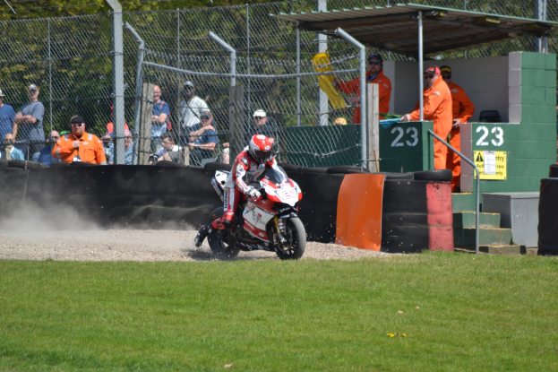 Ein Motorradfahrer mit Helm und Handschuhen fährt über ein grasbewachsenes Feld, mit Zuschauern in Kappen und Schutzbrillen, einem Zaun, Treppen mit Geländern und einem nummerierten Schuppen im Hintergrund.