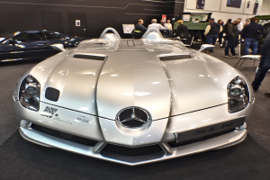 Ein Mercedes-Benz SLR McLaren Roadster wird auf dem Genfer Auto-Salon präsentiert, umgeben von mehreren Bewunderern, während die Ausstellungswände mit Plakaten und Lichtern geschmückt sind.