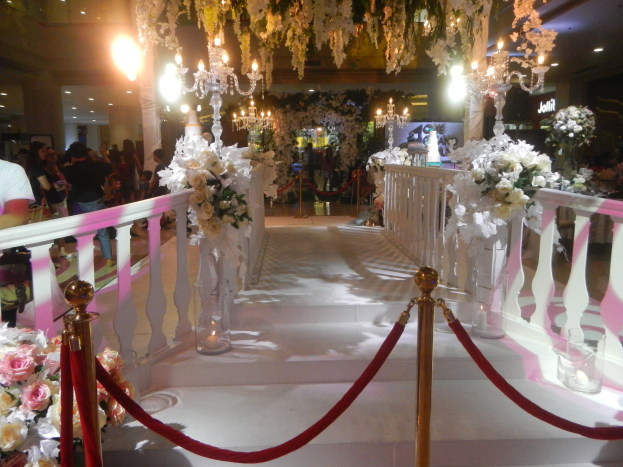 Eine Hochzeitszeremonie in einem Einkaufszentrum mit weißem Teppich, dekorativen Geländern, Blumenarrangements, Kronleuchtern und Deckenbeleuchtung, bei der eine Menge anwesend ist, die Taschen hält.