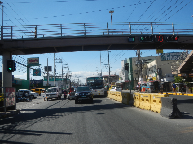 Eine belebte Stadtstraße mit verschiedenen Fahrzeugen, einschließlich Autos, Bussen und Motorrädern, eine Brücke mit Geländern und Laternen, Strommasten mit Drähten und Schilder mit Text unter einem bewölkten Himmel.