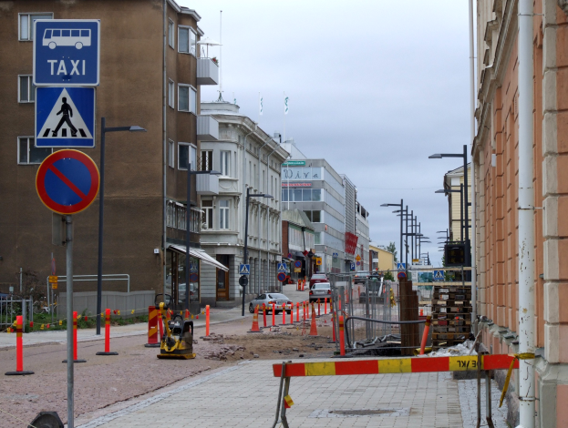 Stadtstraße mit Gebäuden, Fahrzeugen, Verkehrsschildern, Baustelle, Bäumen und Himmel mit Wolken.