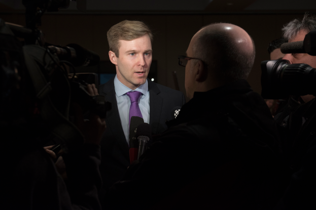 Ein Mann im Anzug und Krawatte, der eine Brille trägt, wird von Reportern interviewt, wobei zwei Personen mit Kameras links und rechts von ihm stehen und ein Mikrofon vor ihm liegt.