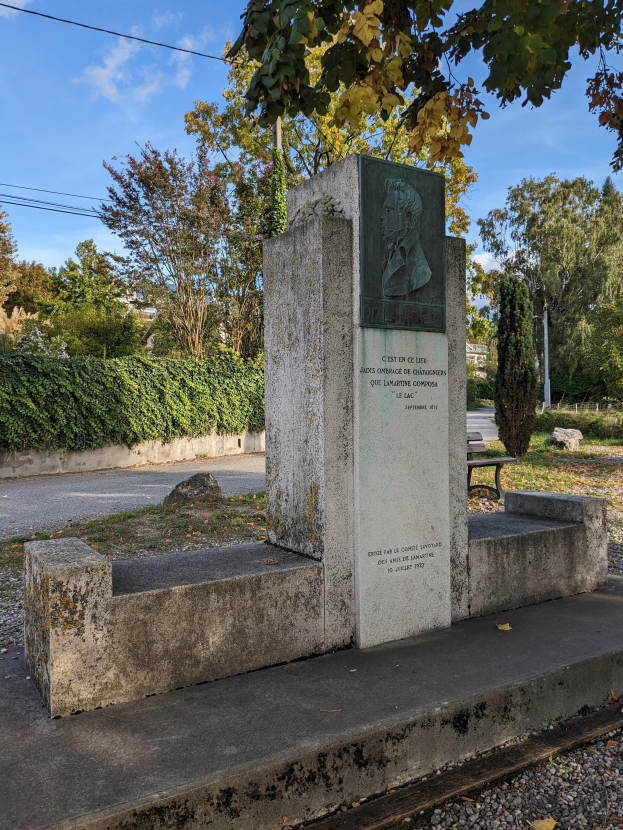 Ein steinernes Denkmal mit eingravierter Schrift steht in einem Park, umgeben von Gras, Pflanzen, Bäumen und einer Bank davor, mit einem bewölkten Himmel darüber.