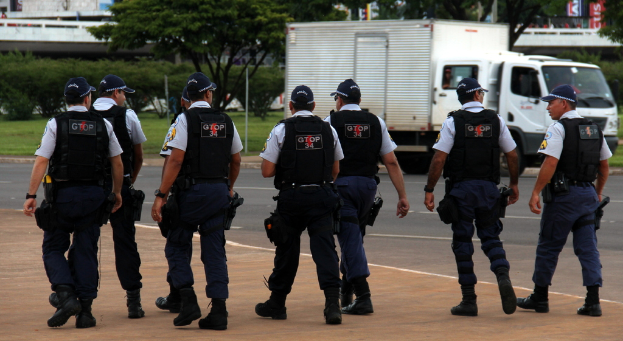Eine Gruppe von Beamten in Uniform, einschließlich Mützen, Hemden, Schutzwesten, Hosen und Schuhen, die auf einer Straße mit einem geparkten Lkw auf der rechten Seite neben einem Zaun, Gebäuden, Bäumen, Pflanzen, Polen und Schildern im Hintergrund und einer Brücke im oberen rechten Eck gehen.