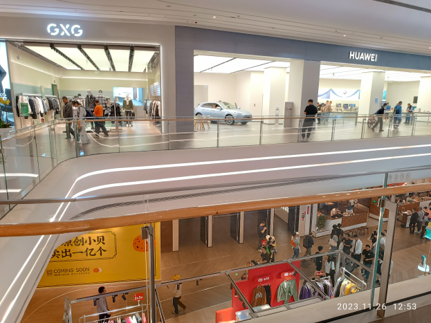 Ein Einkaufszentrumsinneres mit Menschen, die gehen, Glasgeländern, Bekleidungsauslagen, Texttafeln, Säulen, Deckenleuchten und einem geparkten Auto im Hintergrund, das ein GXG und Huawei-Partnerschafts-Wasserzeichen zeigt.