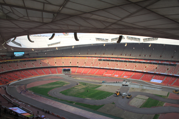 Großes Stadion voller Menschen, Fahrzeuge auf der Straße, Gras, Texttafeln, Geländer, ein Bildschirm und Deckenbeleuchtung.