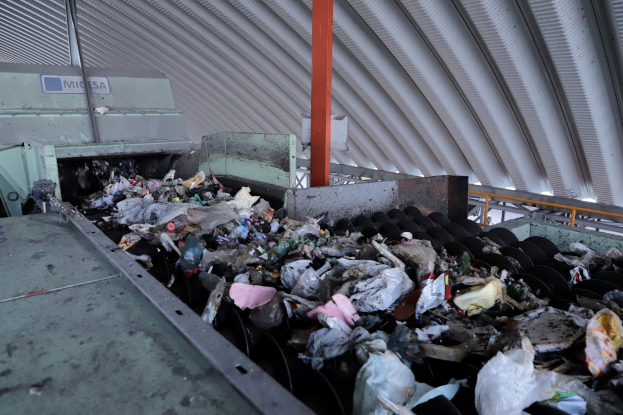 Großer Müllhaufen auf einem Förderband in einer Recyclinganlage mit verstreuter Müll und einem Metalldach.