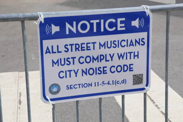 Anschlagtafel an einem Zaun mit einem Schild, auf dem "Anschlag: Alle Straßenmusiker müssen sich an die Stadtlärmvorschriften halten" steht, und einer Straße im Hintergrund.