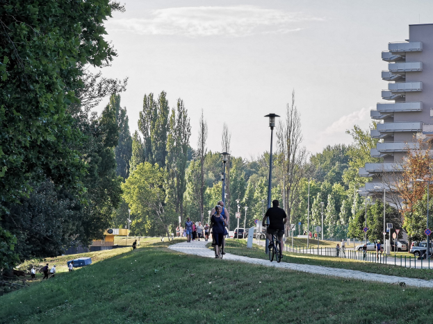 Eine Gruppe von Menschen auf Fahrrädern auf einem Parkweg mit Bäumen, Straßenlaternen, Verkehrsschildern, Kraftfahrzeugen auf der Straße, Absperrpoller, Gebäuden und einem bewölkten Himmel im Hintergrund.