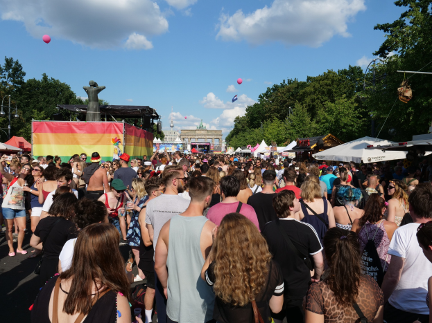 Eine große Menschenmenge marschiert auf einer Straße mit Zelten, Bäumen, Pfählen und Lichtern, mit einer Statue und Gebäuden im Hintergrund, unter einem bewölkten Himmel mit Ballons, während des Berlin Pride Festivals.