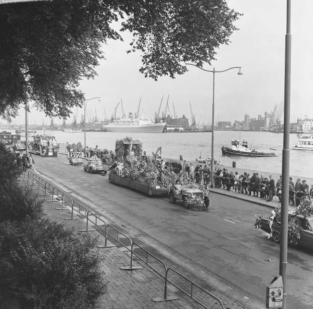 Ein Schwarz-Weiß-Foto einer Parade auf einer Stadtstraße, mit Fahrzeugen auf der Straße, Menschen auf dem Gehweg, Geländern, Pfählen, Laternen, Bäumen, Schiffen auf dem Wasser, Gebäuden und einem Himmel im Hintergrund.
