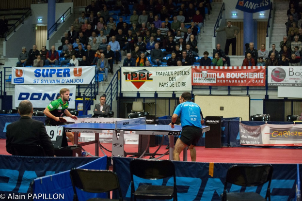 Zwei Männer spielen Tischtennis drinnen, mit einigen Zuschauern, die stehen und sitzen, sowie leeren Stühlen und Werbeplakaten.