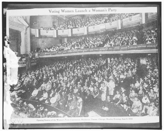 Schwarzes und weisses Foto einer grossen Menge in einem Gebaude mit einigen sitzenden und stehenden Personen, Schilder mit Text, eine Wand im Hintergrund und der Text 'Wahlberechtigte Frauen gruenden eine Frauenpartei' unten.