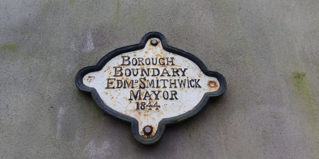 Metallplakette an der Seite eines Gebäudes mit schwarzem Hintergrund und weißer, fetter, zentrierter Schrift, die "Gemeindegrenze Edm Smithwick Bürgermeister" lautet.
