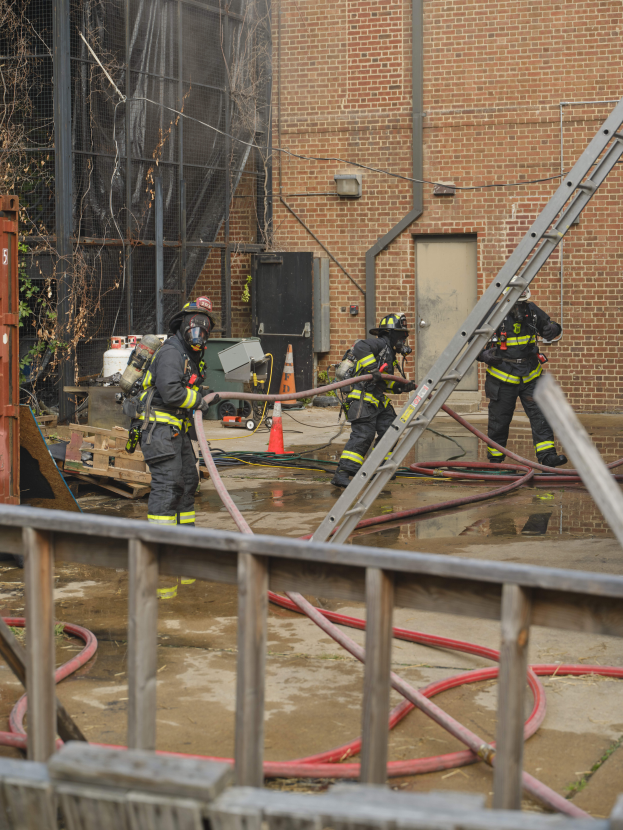 Eine Gruppe von Feuerwehrmännern in Helmen, die Rohre halten, arbeiten daran, ein Gebäude Feuer zu löschen, mit verschiedenen Ausrüstungen und Strukturen drumherum, einschließlich eines Metallzauns, Rohre auf dem Boden, einem Container, einem Verkehrskegel und einem Baum im Hintergrund.