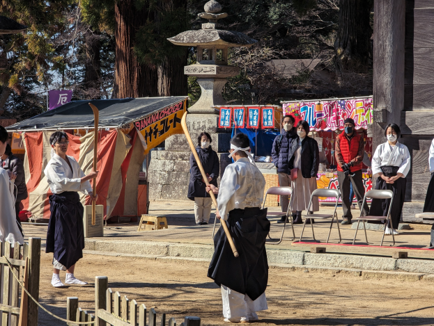 Eine Gruppe von Menschen in formeller Kleidung, einige mit Masken und Stöcken, versammelt sich im Freien vor einem Gebäude während einer traditionellen japanischen Zeremonie in Kyoto, mit Stühlen, einem Zelt, Bannern, einem Pfahl, Bäumen, einem Holzzaun und einem Seil gegen einen klaren blauen Himmel.