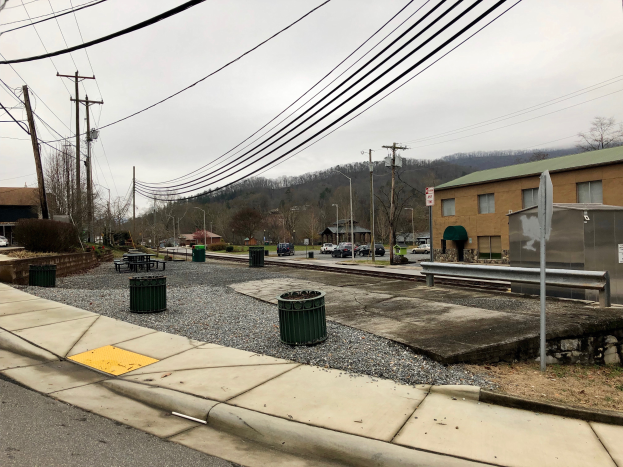 Eine Straßenszene in der Stadt mit einem Bürgersteig, Mülltonnen, Straßenlaterne, Strommasten mit Kabeln, Fahrzeugen, Gebäuden mit Fenstern, Bäumen und einem bewölkten Himmel.
