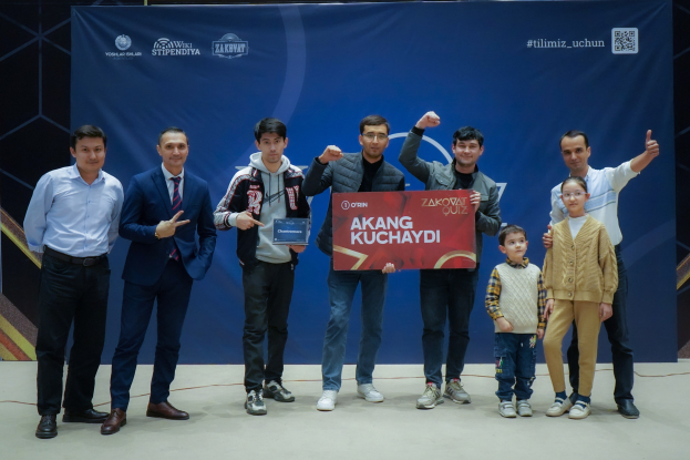 Gruppe von Menschen auf einer Bühne mit einer Tafel, auf der 'Akang Kuchaydi - Zakovat Quiz' steht, mit einem Banner, Lichtern und anderen Gegenständen im Hintergrund.