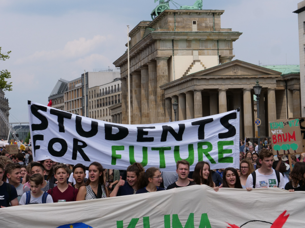 Gruppe von Schülern marschiert in Berlin mit einem leuchtend bunten "Schüler*innen für die Zukunft"-Schild vor einer Kulisse aus Gebäuden, Bäumen und Himmel.