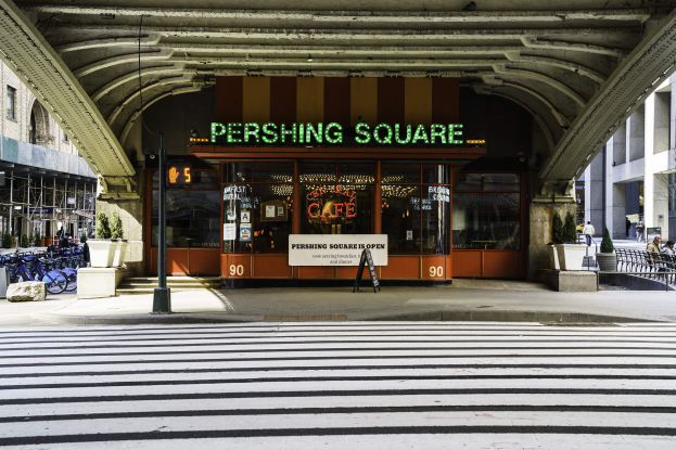 Eine belebte Stadtkulisse mit einem zentralen Gebäude, einem Schild mit der Aufschrift "Pershing Square Cafe in New York City", abgestellten Fahrrädern, Topfpflanzen, einer Straßenlaterne, einem Metallzaun, Menschen auf Bänken sitzend, Bäume und ein bewölkter Himmel.