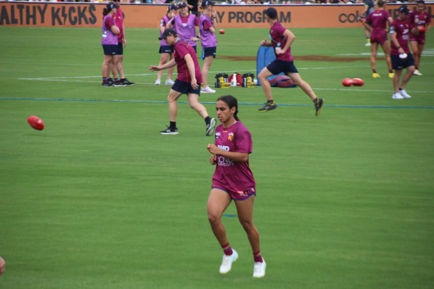 Frauen spielen Australian Rules Football auf einem grünen Feld mit verstreuten Bällen und einem Banner im Hintergrund, einige tragen Kappen und Turnschuhe.