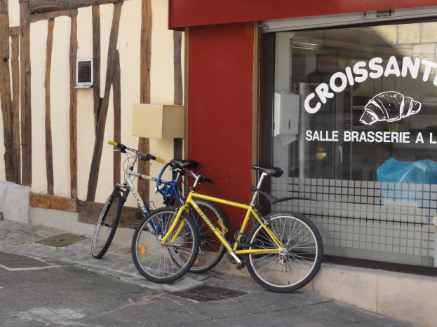 Zwei Fahrräder nebeneinander auf einem Gehweg vor einem Laden mit einer Glaswand und Text, und einem Objekt an der Wand links von den Fahrrädern.