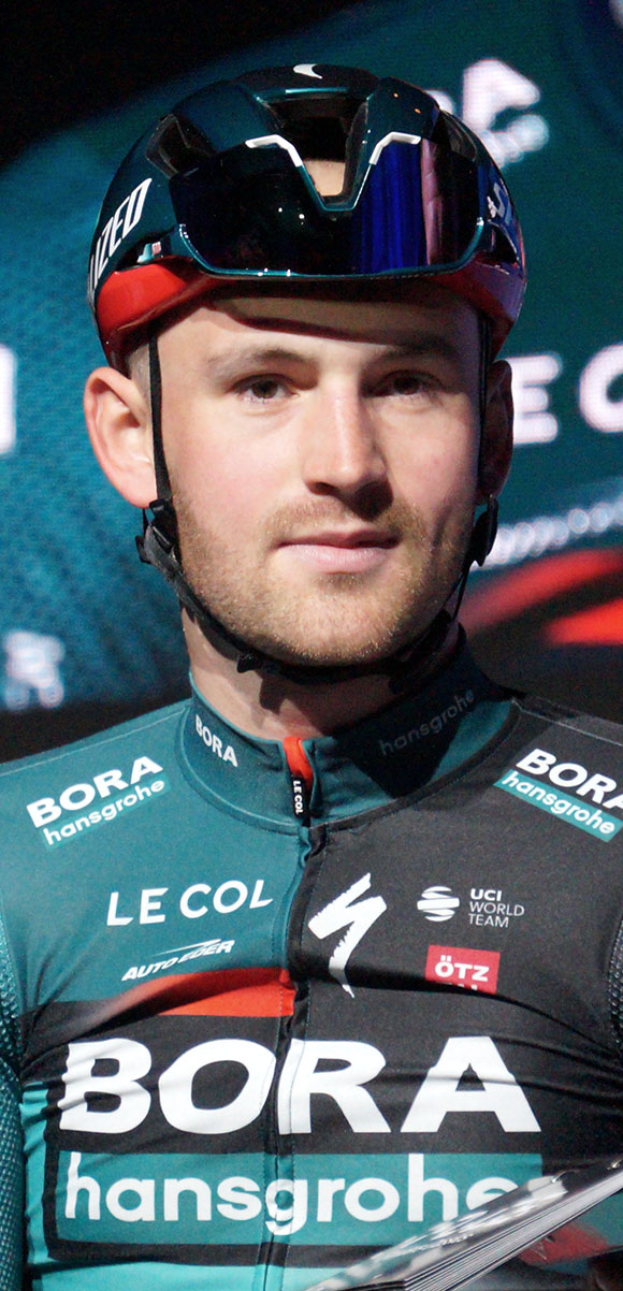Ein Mann in einem blauen Helm und einem Teamtrikot steht neben einem Fahrrad, mit einem 'Bora-Hansgrohe World Tour'-Plakat im Hintergrund und sieht entschlossen aus.