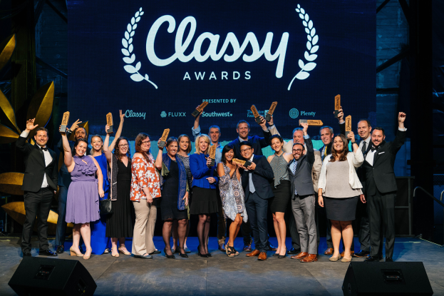Eine Gruppe von Menschen auf einer Bühne, die strahlen und Auszeichnungen halten, mit Lautsprechern unten und einem Hintergrundbild mit "Classy Awards 2019".