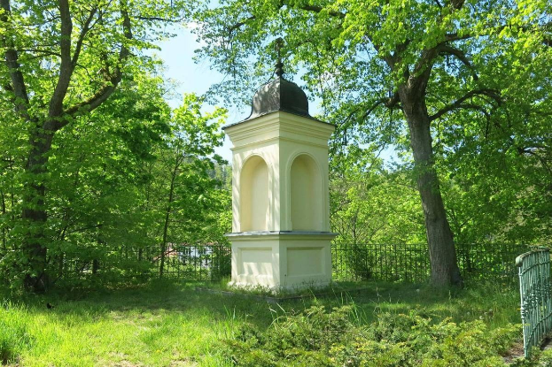Kleines weißes Holocaust-Denkmal auf einem Rasenfriedhof umgeben von einem Zaun und Bäumen unter einem klaren blauen Himmel in Vilnius, Litauen.